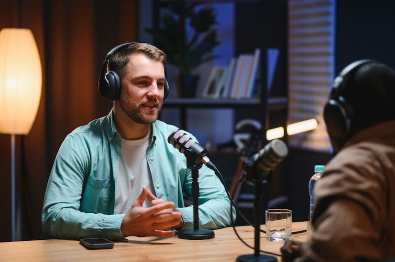 Two people wearing headphones sit at a table with microphones, recording a podcast or interview on reputation management tips in a cozy, dimly lit studio with bookshelves and a floor lamp in the background.