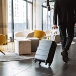 A person in a suit walks through a modern hotel lobby focused on hotel reputation management, pulling a black suitcase, with contemporary yellow and gray chairs and large windows in the background.