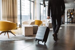 A person in a suit walks through a modern hotel lobby focused on hotel reputation management, pulling a black suitcase, with contemporary yellow and gray chairs and large windows in the background.
