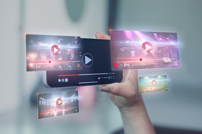 A hand holds a smartphone displaying multiple floating video play screens with progress bars and play buttons, symbolizing streaming or virtual media content—hinting at what is the Streisand effect when online videos go viral, against a blurred background.