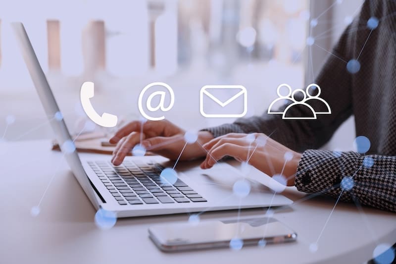 A person types on a laptop at a desk with a smartphone nearby. Transparent icons of a phone, email, and people are overlaid, hinting at communication options—much like what is Radaris used for: finding contact details and connecting with others.