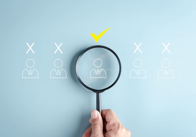A hand holds a magnifying glass over one of four illustrated figures in suits, highlighting the process of selection—similar to what is Radaris used for when verifying identities. The chosen figure has a yellow checkmark; others have white X marks.