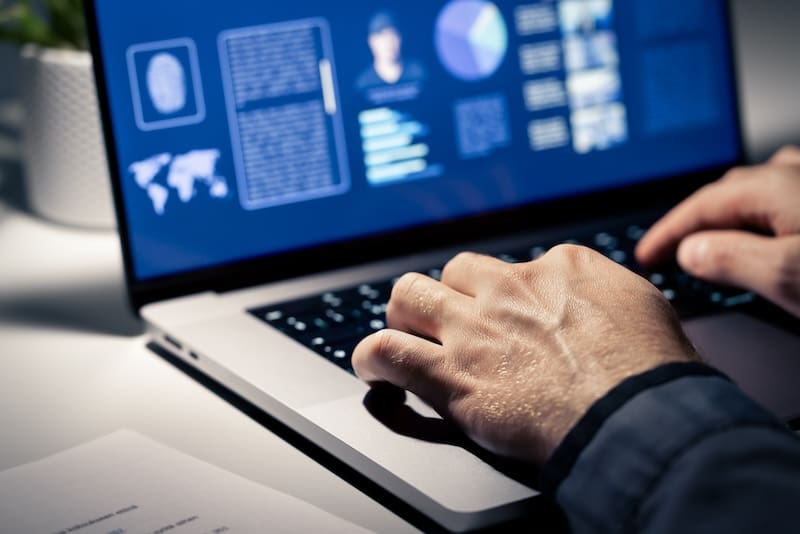 A person’s hands typing on a laptop keyboard, with a screen displaying various data, charts, and graphics in dark mode—perhaps researching what Radaris is used for. A sheet of paper and a plant are partially visible on the desk.