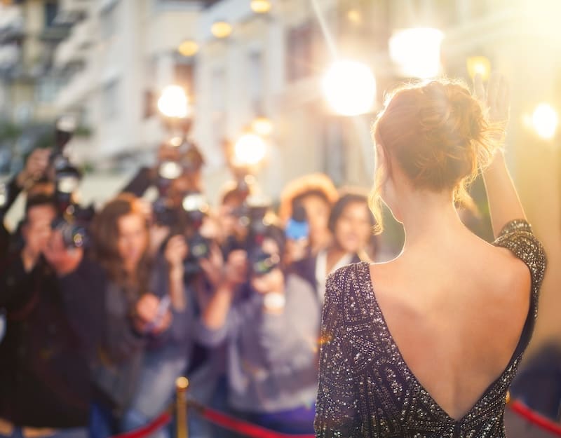A woman in a sparkling dress stands on a red carpet, facing a crowd of photographers with flashing cameras, expertly waving—a true display of celebrity reputation management amid the blur of bright lights in the background.