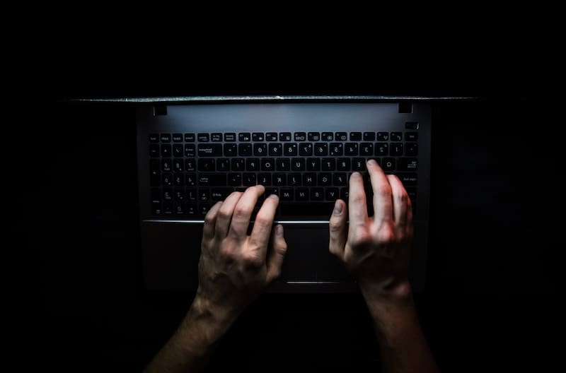 A person’s hands typing on a laptop keyboard in a dark environment, with the screen casting a faint light—suggesting the secretive feel of a dark web photo search.