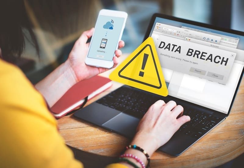 A person uses a smartphone and laptop, both showing warning signs. The laptop screen displays a large "DATA BREACH" alert with an exclamation mark, hinting at threats like dark web photo search and other security issues.