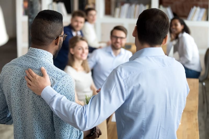 Two men stand facing a group of smiling colleagues around a conference table in an office, one extending his arm around the other's shoulder, suggesting a warm welcome or introduction and highlighting strong job presence.