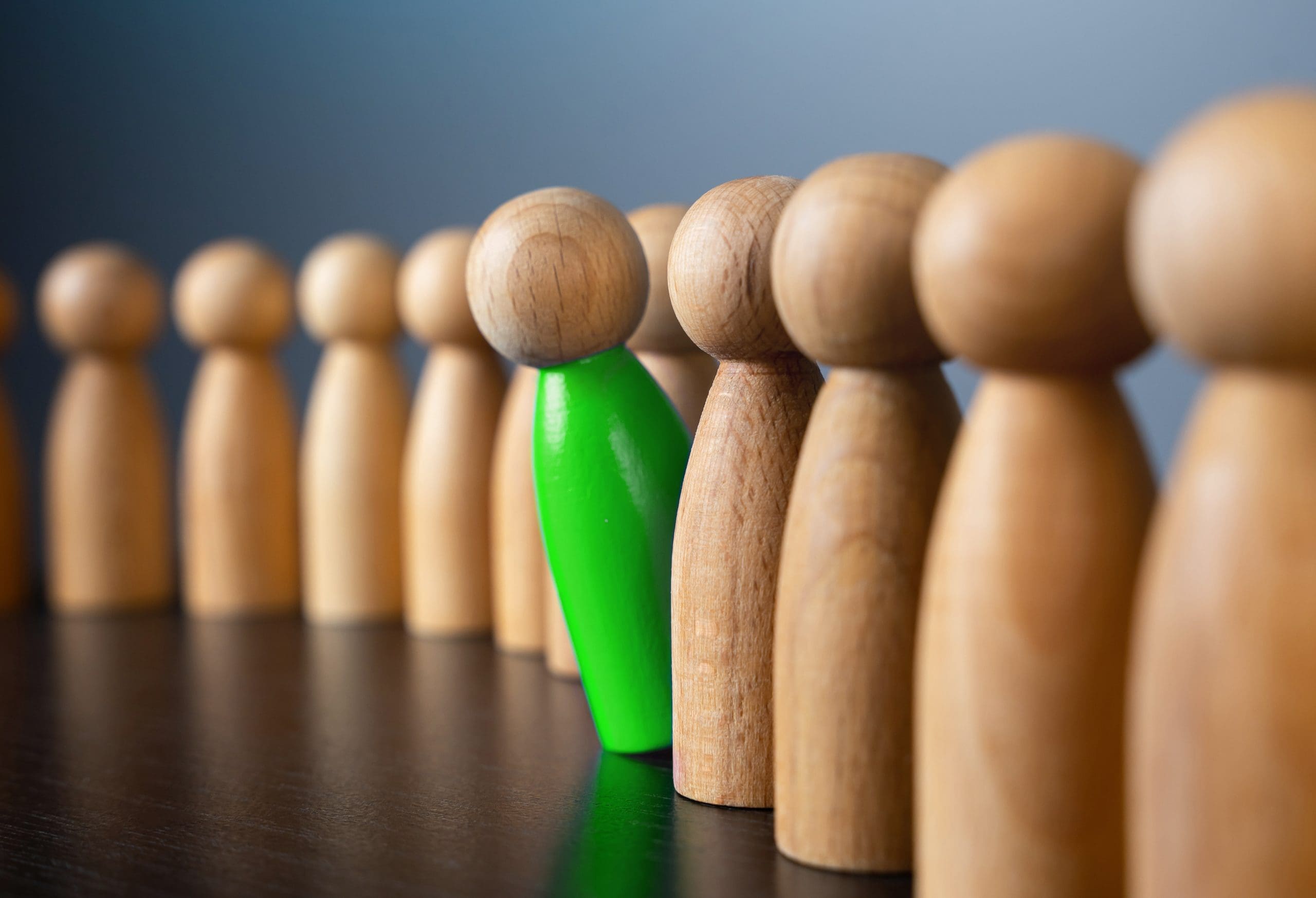 A row of wooden figurines stands on a dark surface, with one green-painted figure among the natural wood-colored ones, symbolizing individuality or standing out from the crowd.