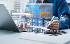 A person working on a laptop and tablet with project management charts, graphs, and reputation tracking tools overlaid digitally, representing planning, scheduling, and data analysis in a modern office setting.