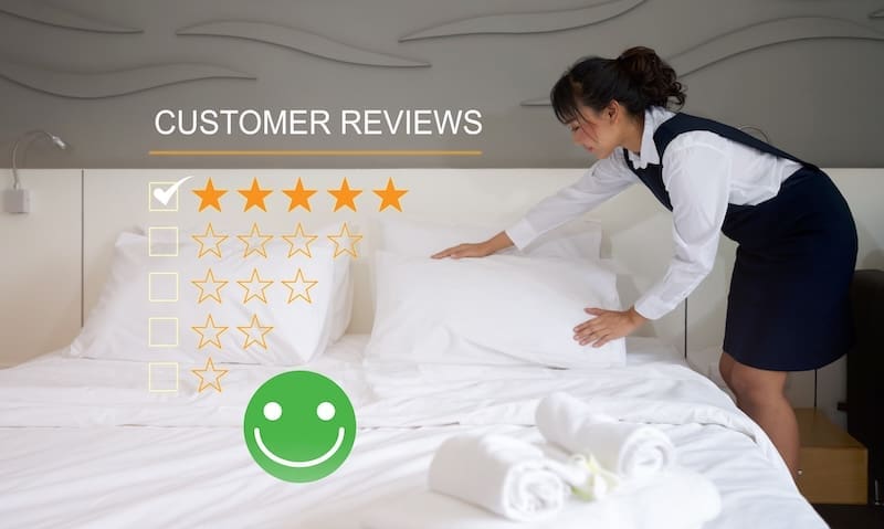 A hotel housekeeper straightens pillows on a neatly made bed, with a five-star customer review rating and a green smiling face graphic, reflecting excellent hotel reputation management.