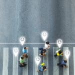 Aerial view of people crossing a street at a crosswalk, with digital location icons above their heads, symbolizing tracking or geolocation technology.