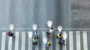 Aerial view of people crossing a street at a crosswalk, with digital location icons above their heads, symbolizing tracking or geolocation technology.