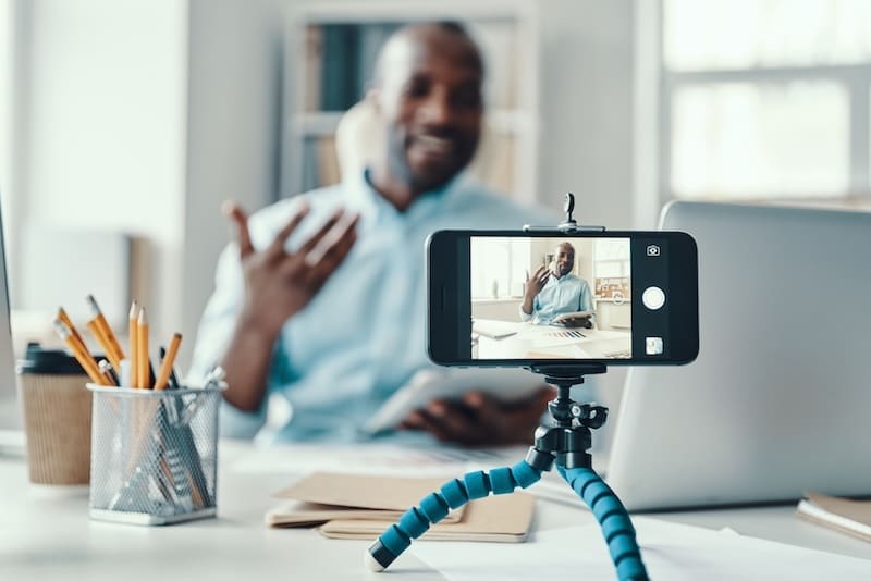 An internet influencer sits at a desk speaking in front of a smartphone mounted on a tripod, recording a video. The desk holds office supplies, papers, and a laptop. The background features a bright, modern office.