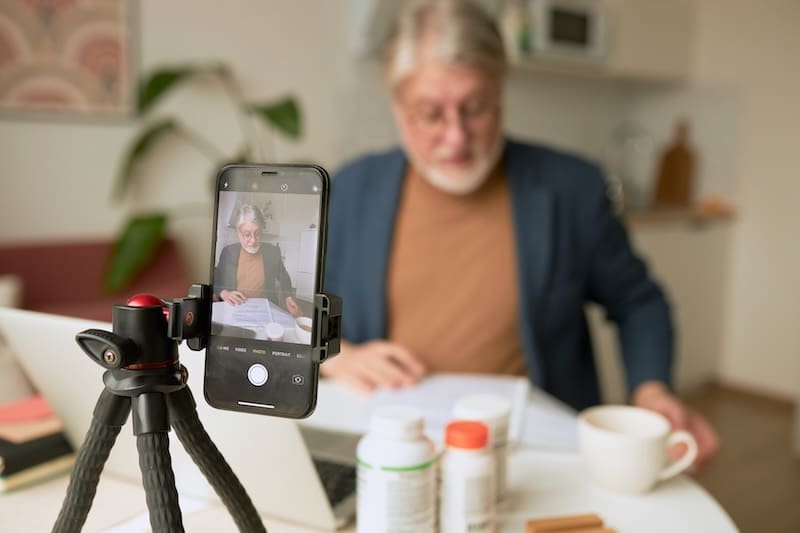 An older man sits at a table with books, bottles, and a cup, while a smartphone on a tripod records him. The phone screen shows his image as he explains something, capturing the look of an internet influencer sharing wisdom online.