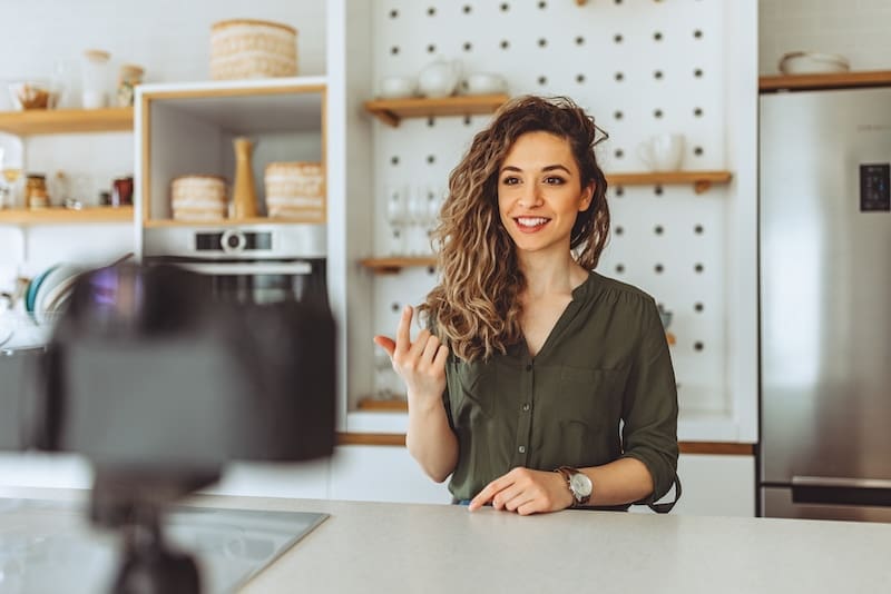 A woman with curly hair and a green shirt sits at a kitchen counter, smiling and gesturing while recording herself on camera as an internet influencer. Shelves and a fridge are visible in the background.