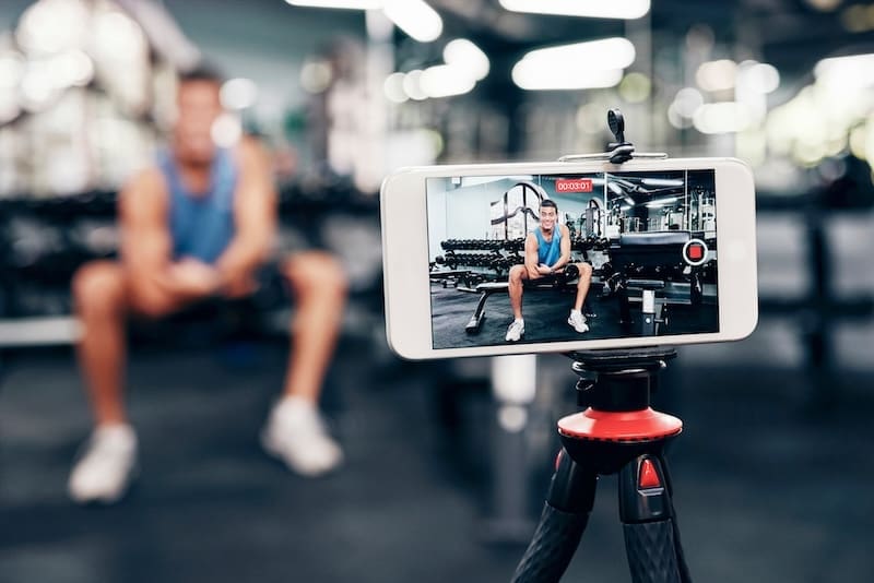 A smartphone on a tripod records an internet influencer in athletic clothing sitting on a bench in a gym, with weights and fitness equipment visible in the background.