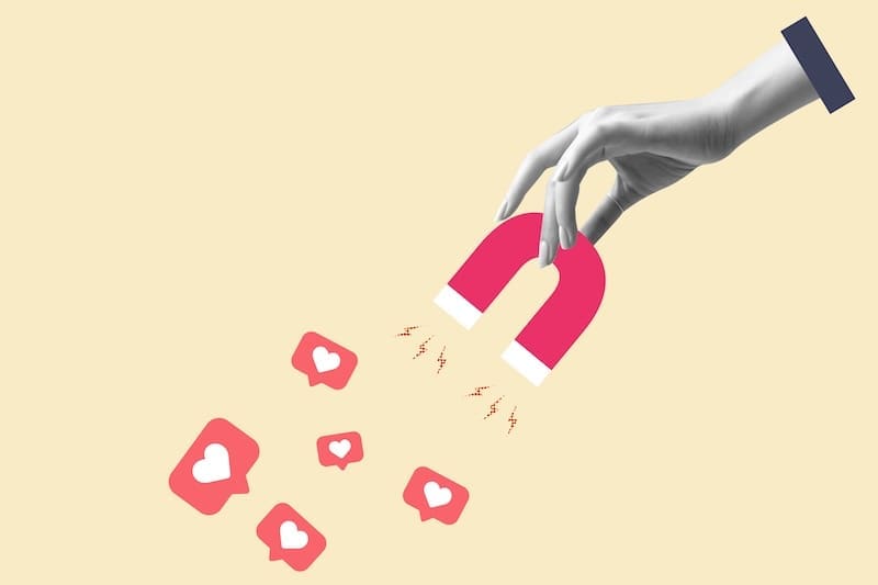 A hand holds a red horseshoe magnet attracting social media like icons with heart symbols, evoking the magnetic pull of an internet influencer, set against a light beige background.