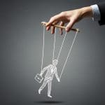 A hand in a suit holds strings attached to a drawn puppet figure of a person carrying a briefcase, symbolizing control or manipulation, against a dark gray background.