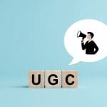 Wooden blocks spelling “UGC” sit on a blue background. Above them is a speech bubble containing an illustrated person holding a megaphone, symbolizing user-generated content or user voices.