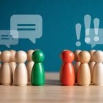 Wooden figures in two groups face each other on a table. One group with a green figure has speech bubbles, while the other group with a red figure has exclamation marks above them, symbolizing a discussion or debate.