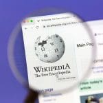 A computer screen shows the Wikipedia main page with the Wikipedia logo magnified by a handheld magnifying glass. The web address bar and part of the page layout are visible.