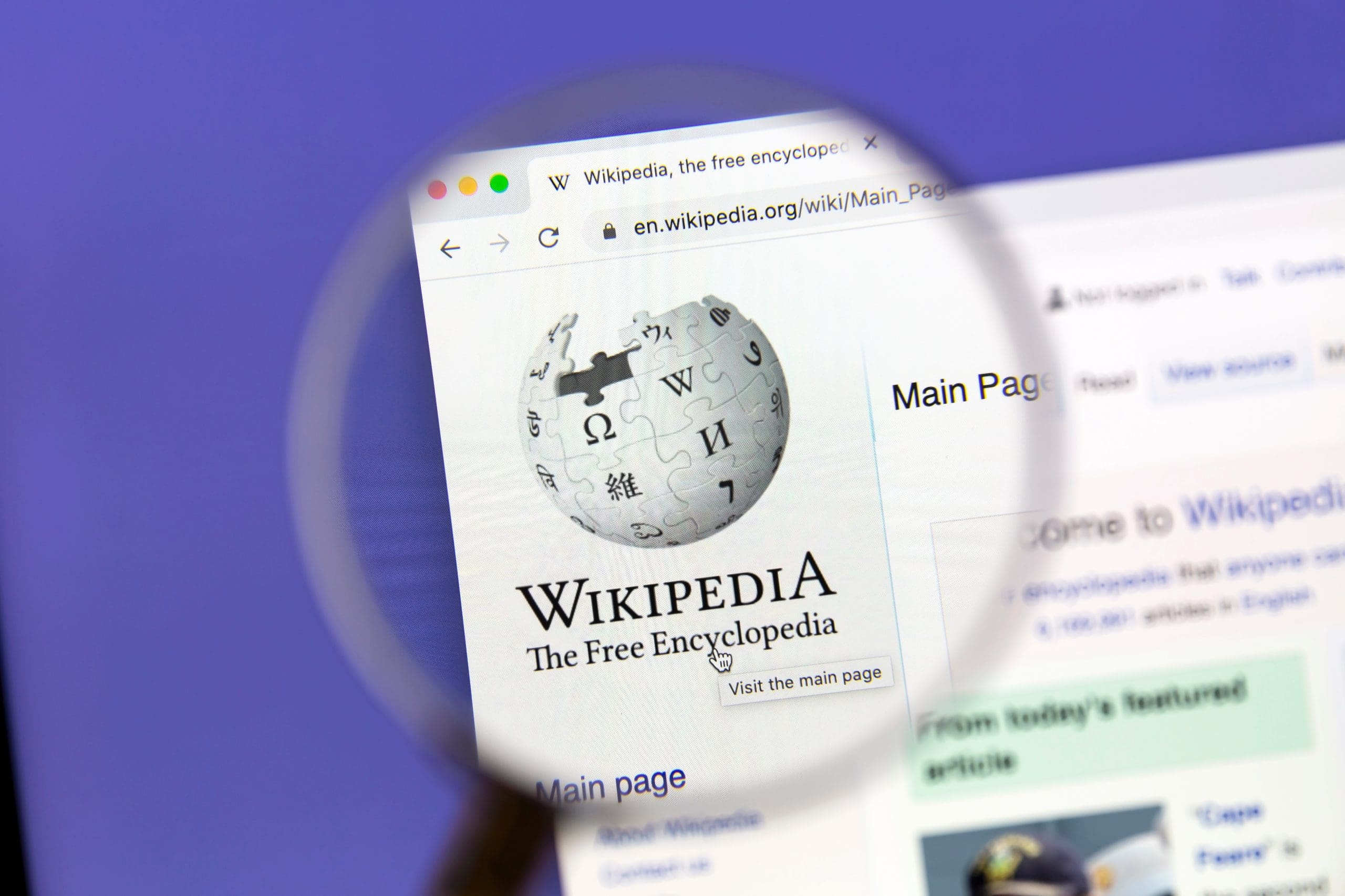 A computer screen shows the Wikipedia main page with the Wikipedia logo magnified by a handheld magnifying glass. The web address bar and part of the page layout are visible.