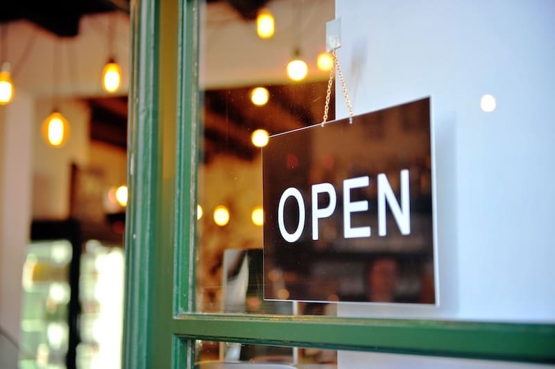A black and white "OPEN" sign hangs on a glass door with a green frame. Warm, blurred lights and shelves hint at the cozy interior, reflecting the welcoming franchise reputation of the shop or café.