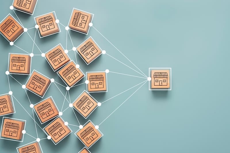 Wooden blocks with storefront illustrations are connected by white lines on a light blue background, symbolizing a strong franchise reputation branching out from a central store to multiple smaller stores.