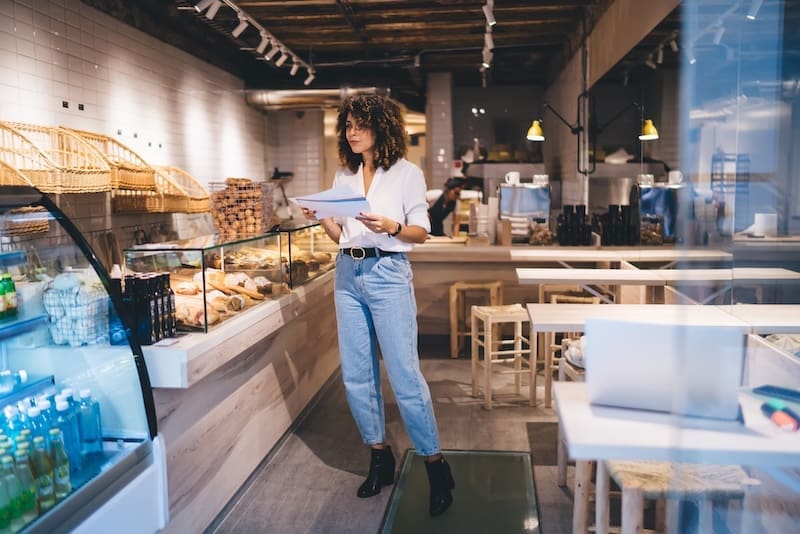 A woman stands in a modern bakery or café, holding papers and looking thoughtful. Surrounded by baked goods, she reflects on the franchise reputation in the bright, stylish interior with wooden tables and a well-stocked display case.