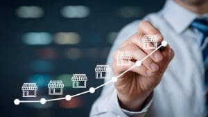 A person in business attire draws an upward-curving graph with storefront icons along the line, symbolizing franchise reputation and business growth. The blurred background suggests an office or corporate environment.