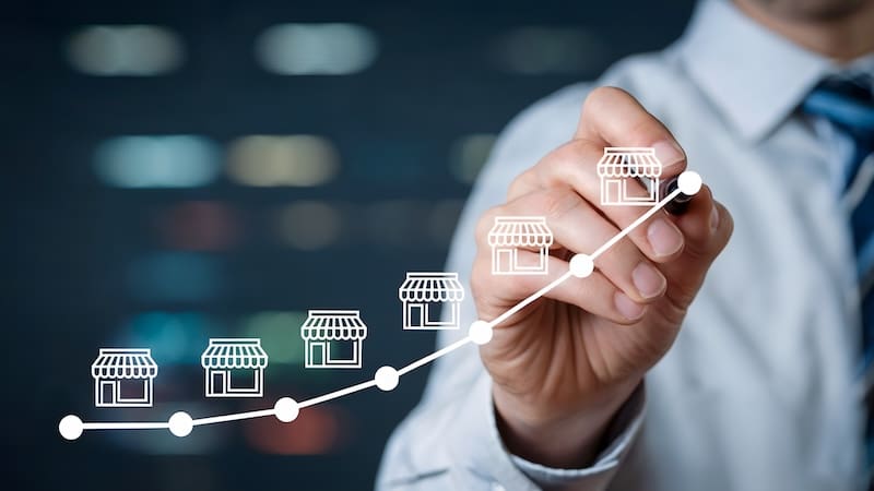 A person in business attire draws an upward-curving graph with storefront icons along the line, symbolizing franchise reputation and business growth. The blurred background suggests an office or corporate environment.