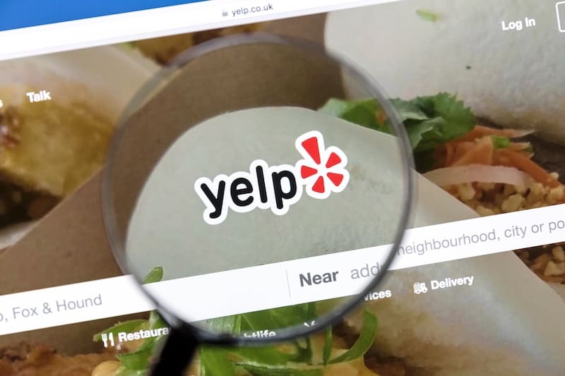 A magnifying glass hovers over a computer screen displaying the Yelp website logo, with blurred images of food and search options visible in the background.