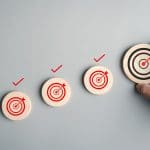 A hand holds a wooden disk with a red target and arrow, placed beside three similar disks, each with a red checkmark above it, symbolizing achieving goals or completing tasks.