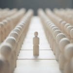 A single wooden figure stands between two rows of identical wooden figures, emphasizing individuality among uniformity.