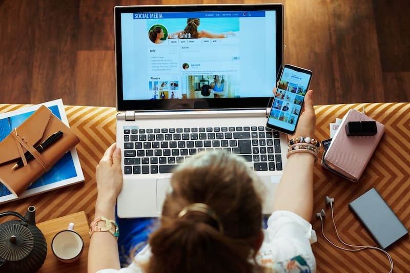 Upper view of young housewife sitting on couch in the modern living room checking social media account on a laptop and smartphone.