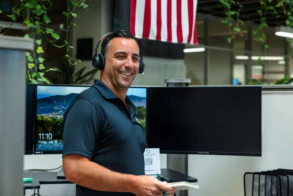 A man wearing a headset and holding a pen smiles while standing in an office with computer monitors. An American flag hangs in the background, and plants are visible nearby.