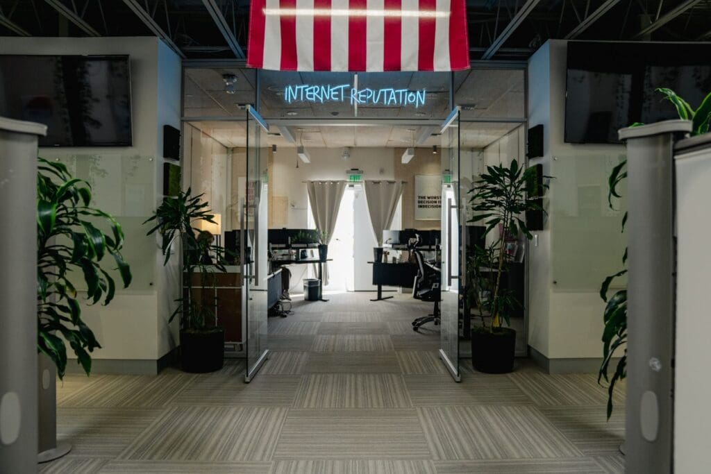Modern office entrance with glass doors, large potted plants on each side, desks and computers visible inside, and a neon sign above reading “INTERNET REPUTATION.” An American flag hangs from the ceiling.