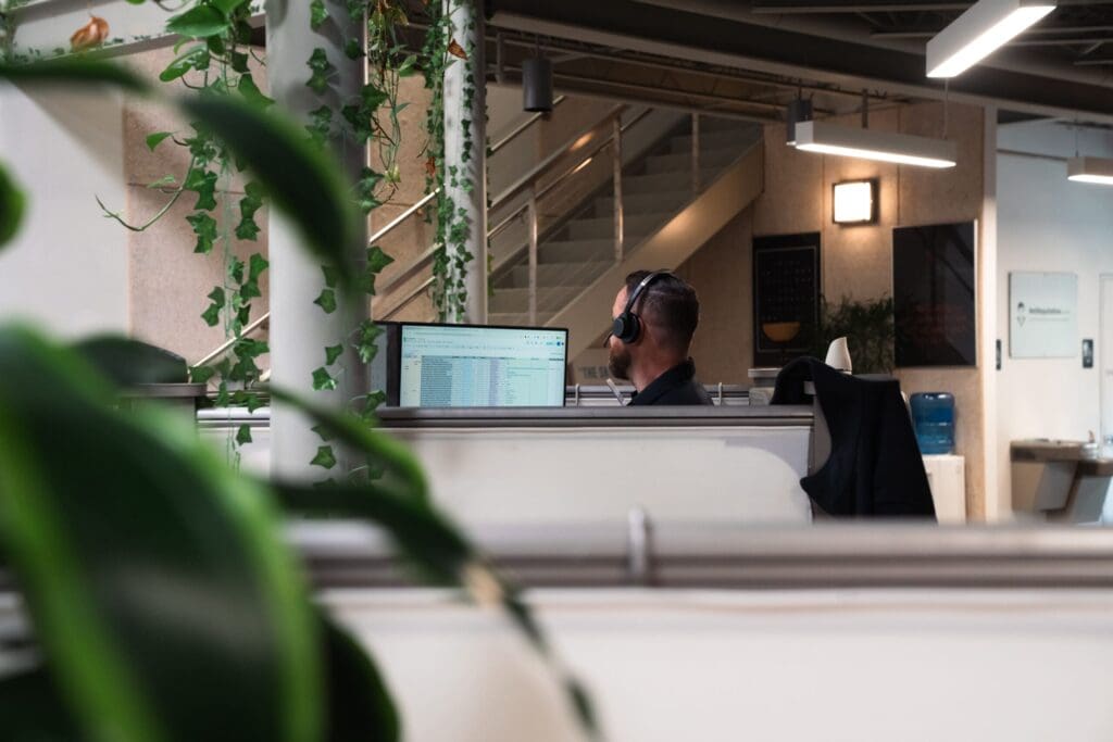 A person wearing headphones sits at a desk in a modern office cubicle, working on a computer. Green plants hang nearby, and a staircase and water cooler are visible in the background.