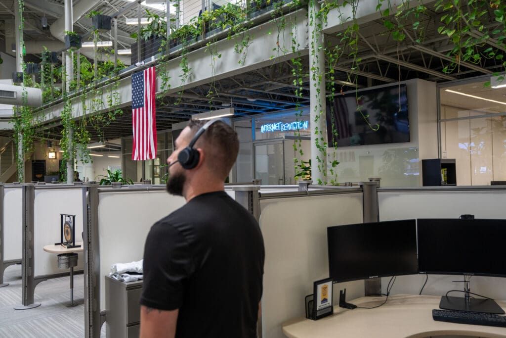 A man wearing a headset stands in a modern office with cubicles, plants, an American flag, and a visible "Internal Revenue" sign in the background. Two computer monitors sit on a nearby desk.